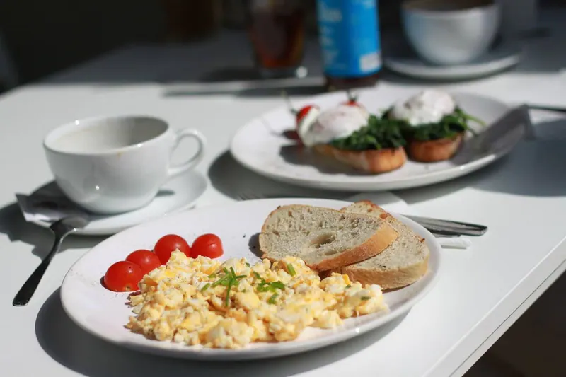 Rührei mit Schnittlauch, Brot und Kirschtomaten auf weißem Teller, daneben Kaffeetasse im Tageslicht